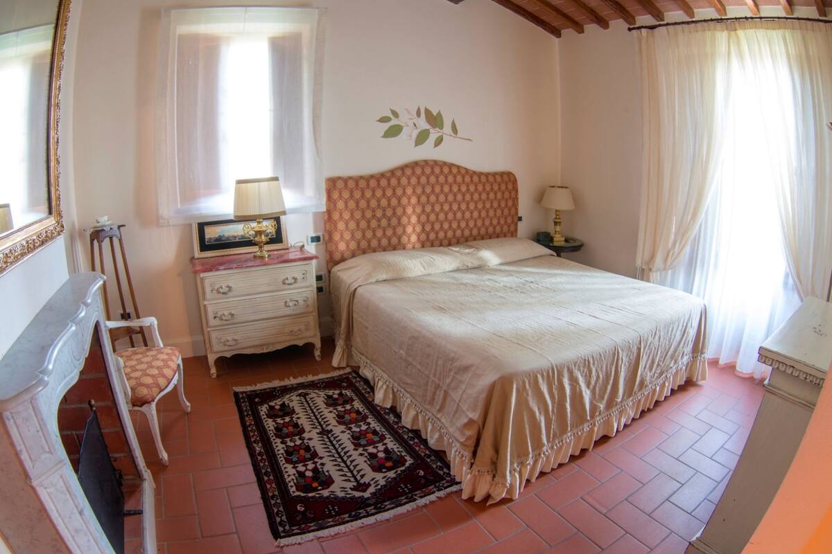 A bedroom with a bed, a nightstand, a chair, a dresser, a window with curtains, and a decorative wall piece.