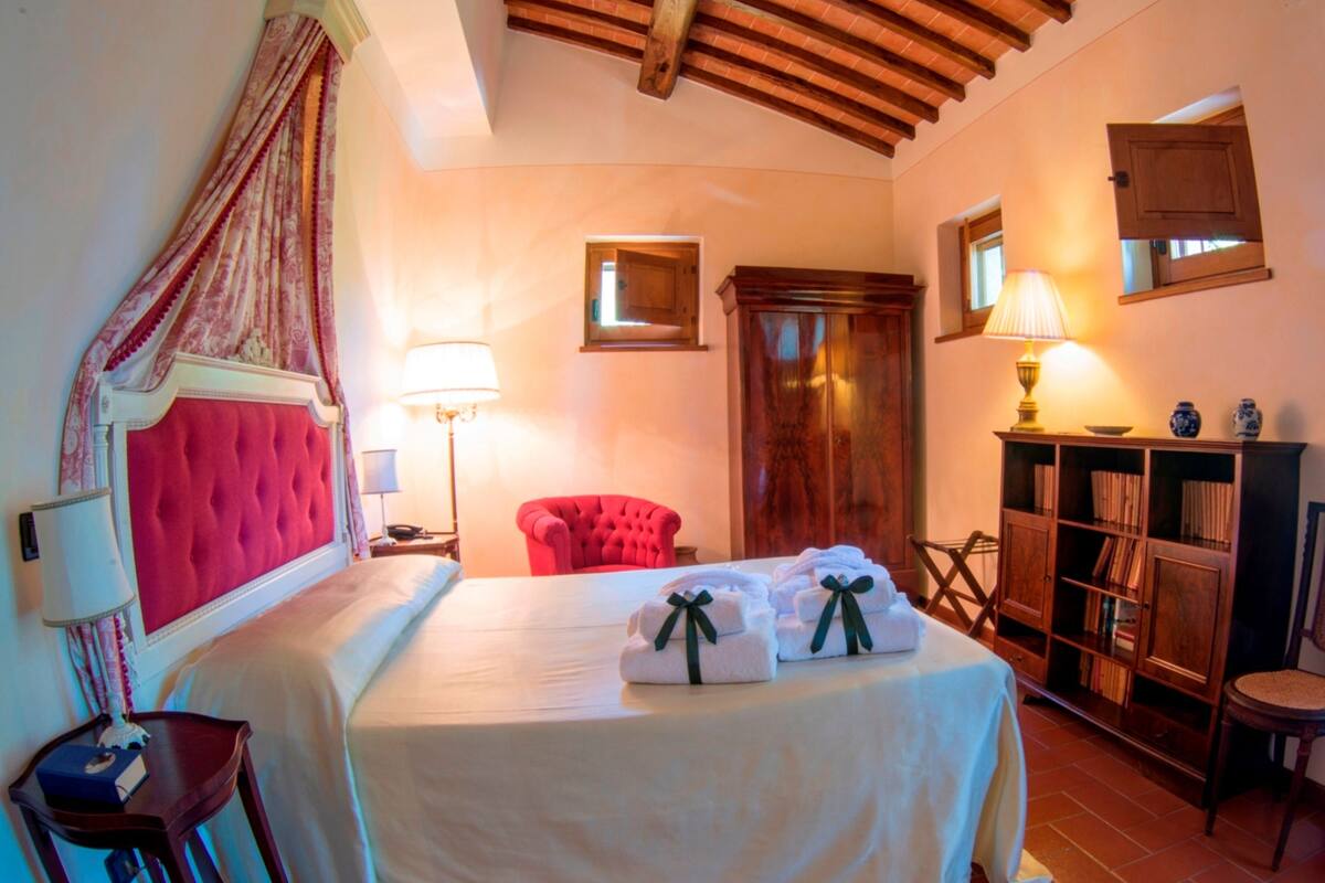 A hotel room with a large bed, a red armchair, a wooden bookshelf, and a small table.