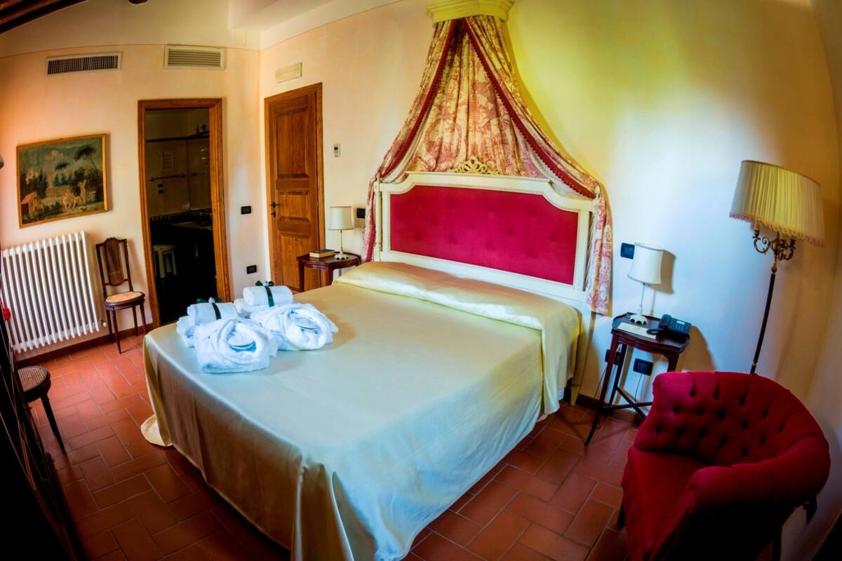 A hotel room with a large bed, a red chair, a radiator, and a painting on the wall.