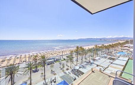 Vista a la playa desde un balcón, con palmeras, piscina y zona de playa con sombrillas.