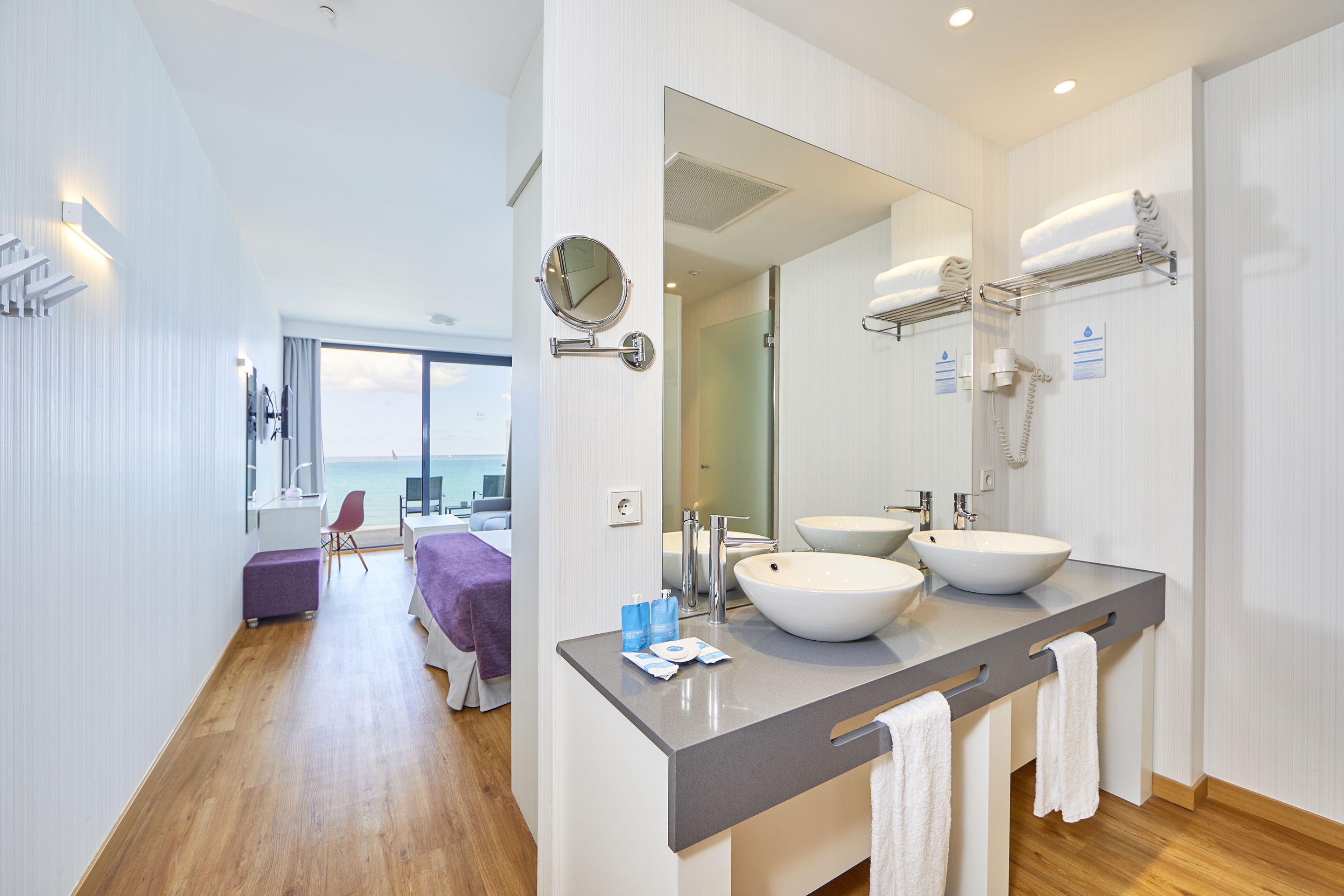 A modern bathroom with two sinks, a large mirror, and a view of the sea through the window.