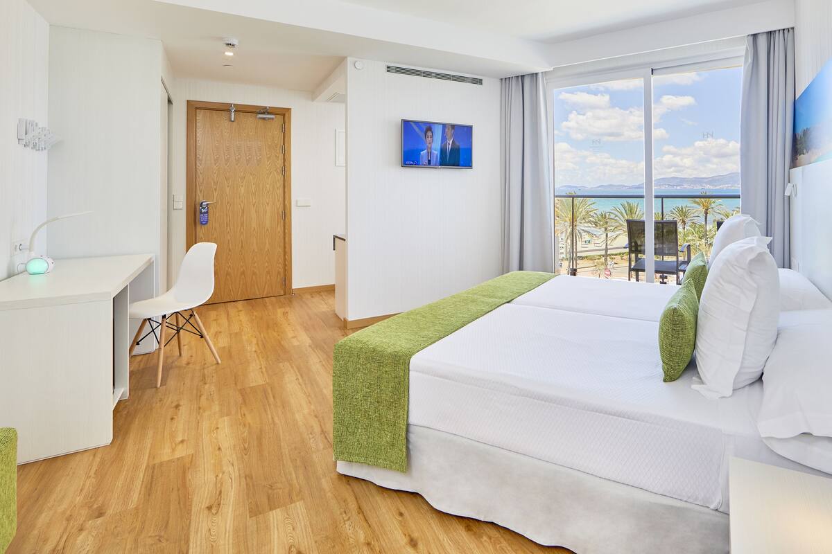 Habitación de hotel con cama, escritorio, silla, televisor y balcón con vista al mar.