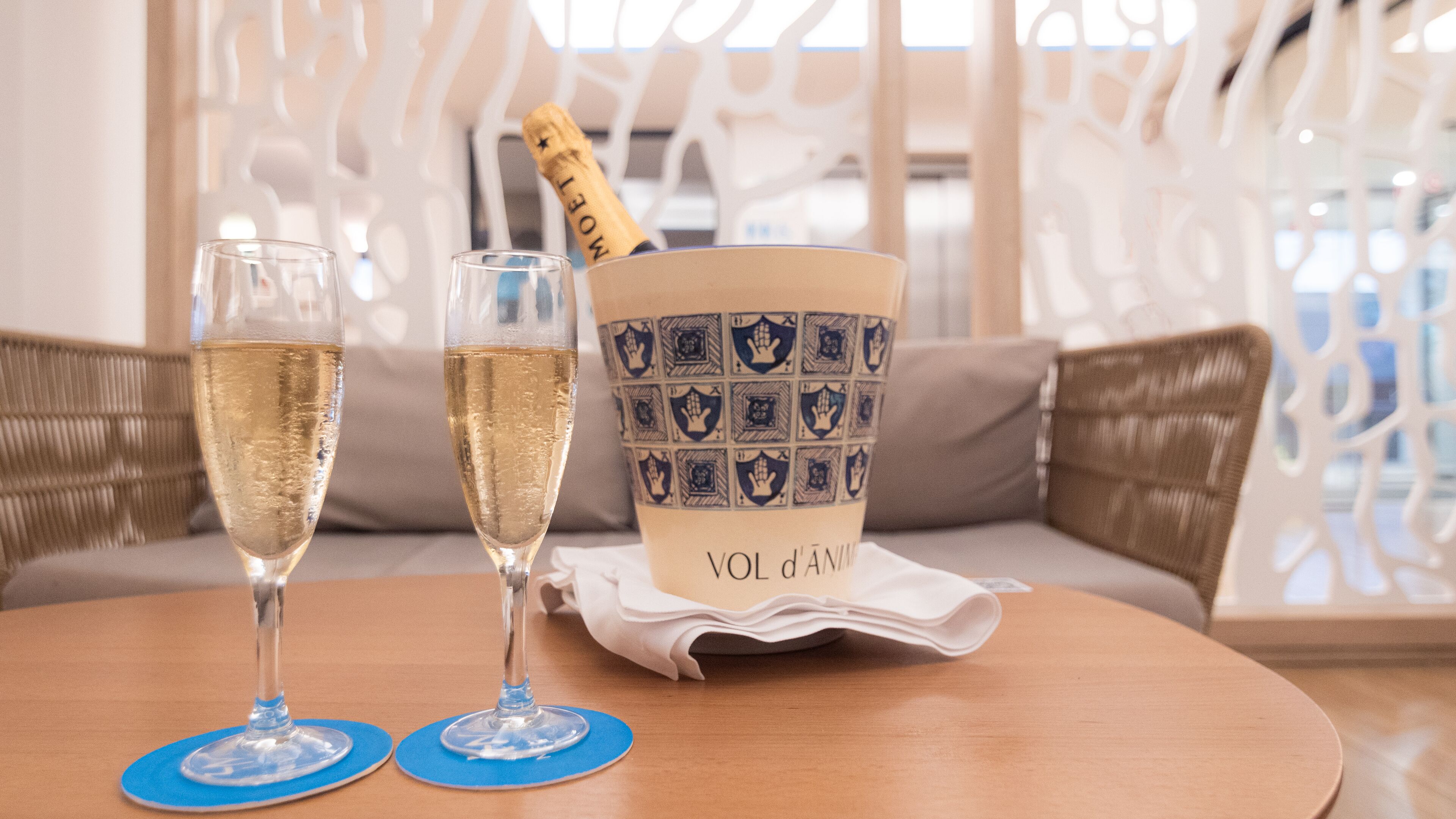 Deux flûtes à champagne posées sur des dessous de verre bleus, un vase décoratif contenant une bouteille de champagne et un coussin sur une table ronde.