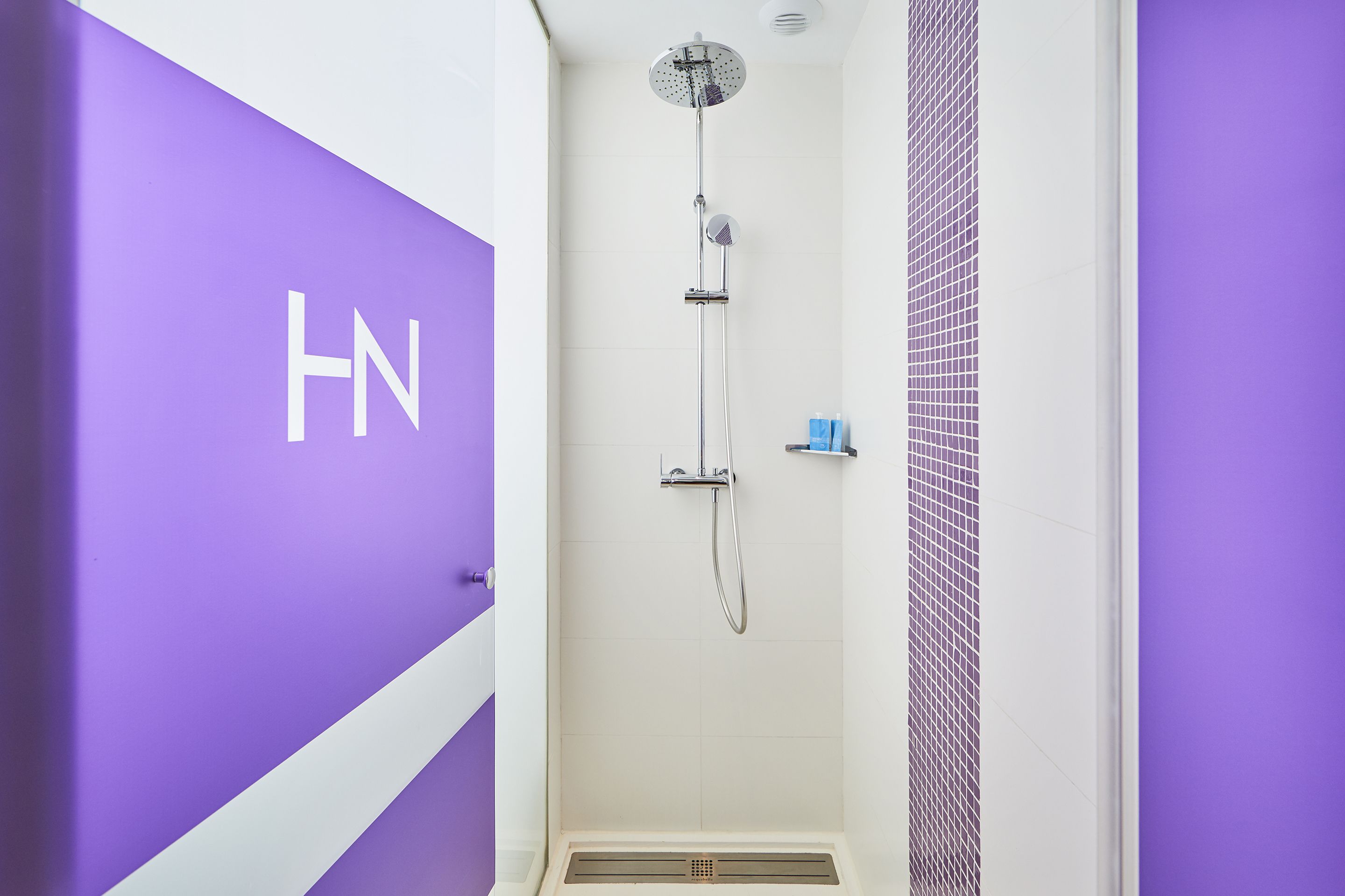Un baño con ducha, pared de azulejos blancos y una pared de acento púrpura con las letras "HN".