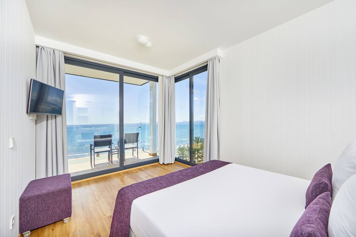Habitación de hotel con una cama grande, un televisor montado en la pared y un balcón con vistas al mar.