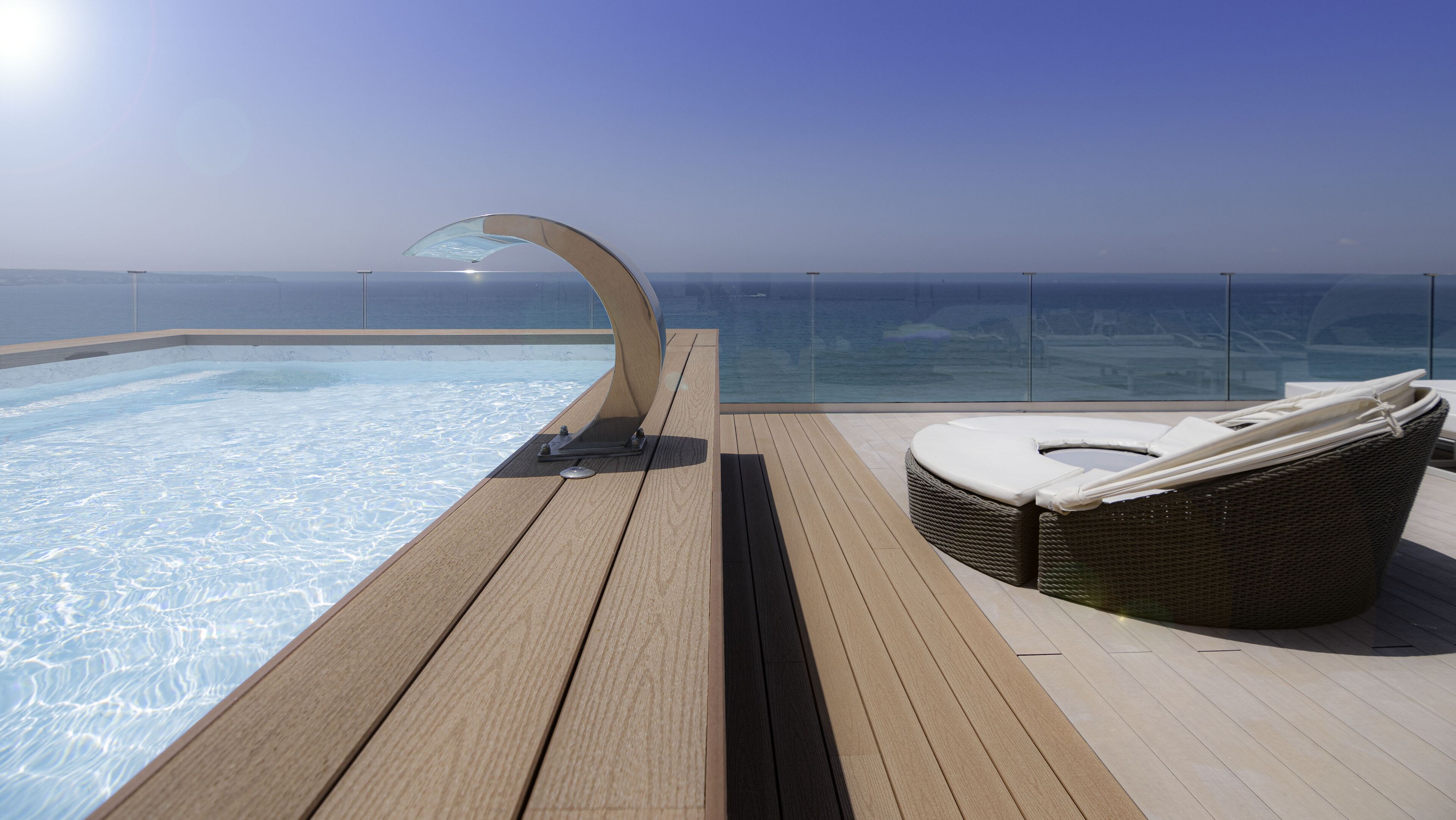 Une piscine extérieure baignée de soleil, dotée d’un jacuzzi à moitié fermé par des parois de verre et d’un fauteuil de relaxation en osier sur une terrasse en bois.