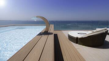 Une piscine extérieure baignée de soleil, dotée d’un jacuzzi à moitié fermé par des parois de verre et d’un fauteuil de relaxation en osier sur une terrasse en bois.