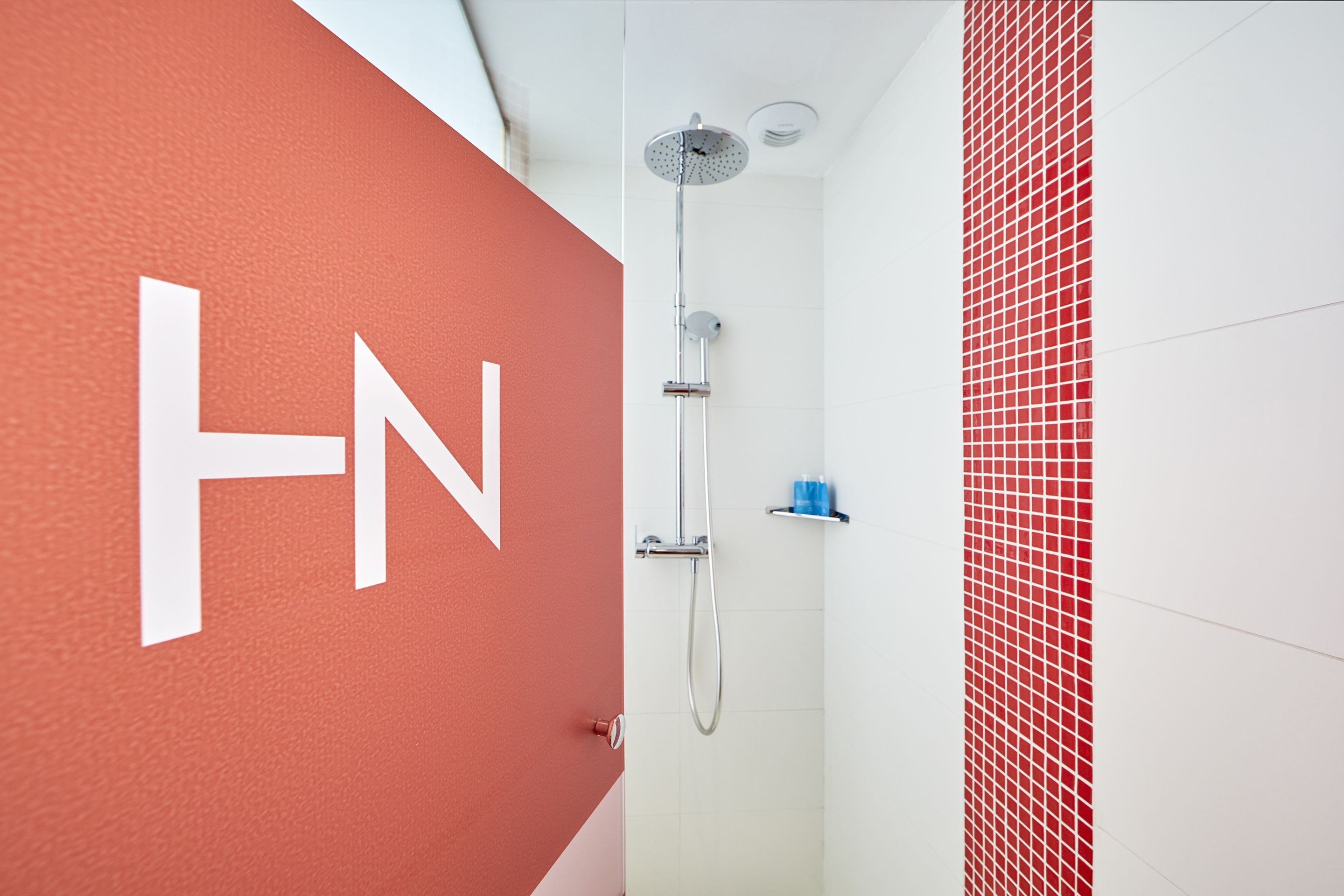 A modern bathroom with a large 'HN' sign on the wall, a shower with a handheld showerhead, and a tiled wall with a red and white pattern.
