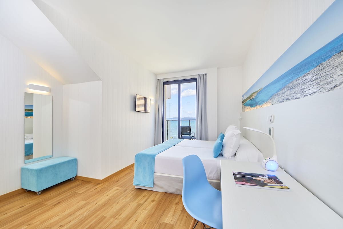 Una habitación de hotel moderna con una cama grande, un escritorio, una silla, un banco y un cuadro de una playa.