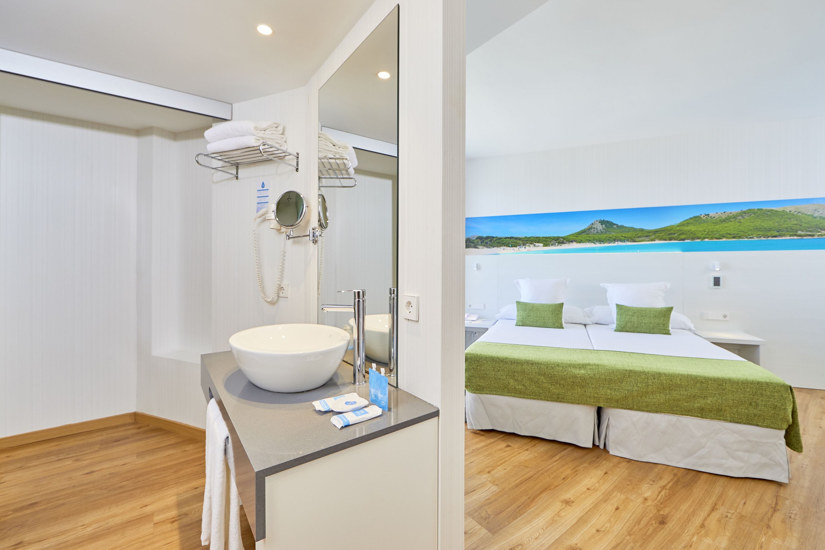 A modern bathroom with a large sink, mirror, and towel rack. The room features a wooden floor and a bed with a green blanket and white pillows.