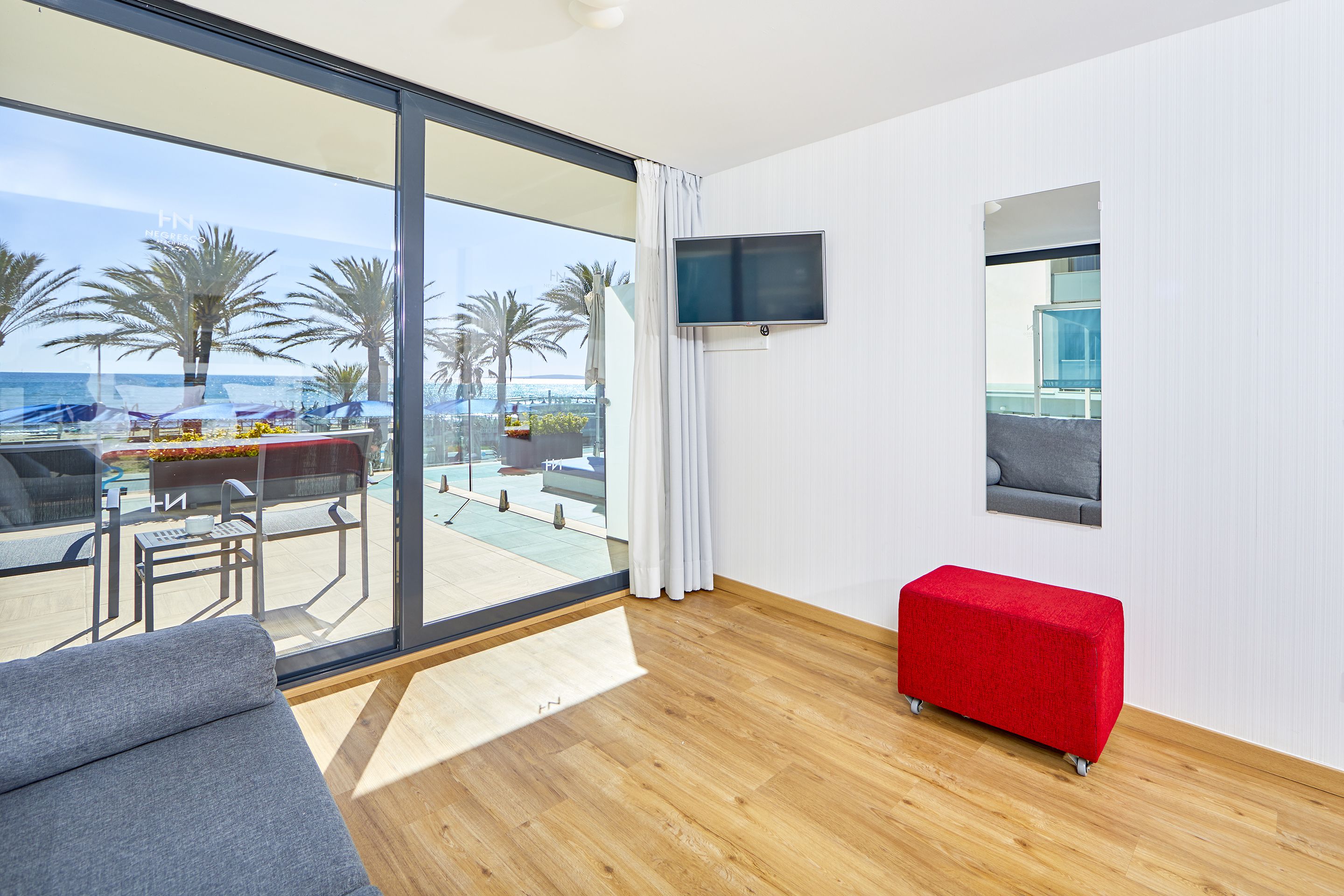 A modern room with a large glass door leading to a balcony with outdoor seating and palm trees.