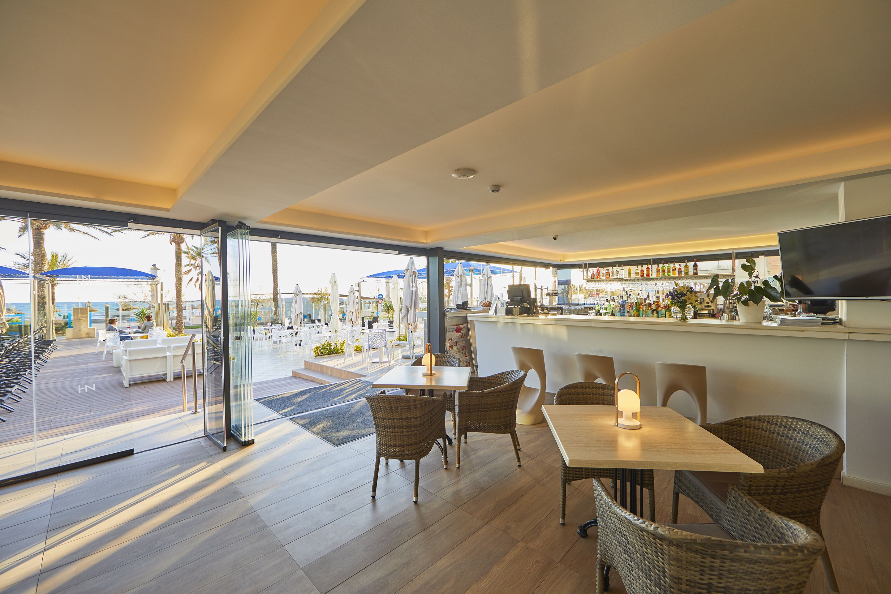 Un comedor interior con sillas de mimbre y una mesa, una barra con televisor y ventanas amplias que ofrecen vista a la playa.