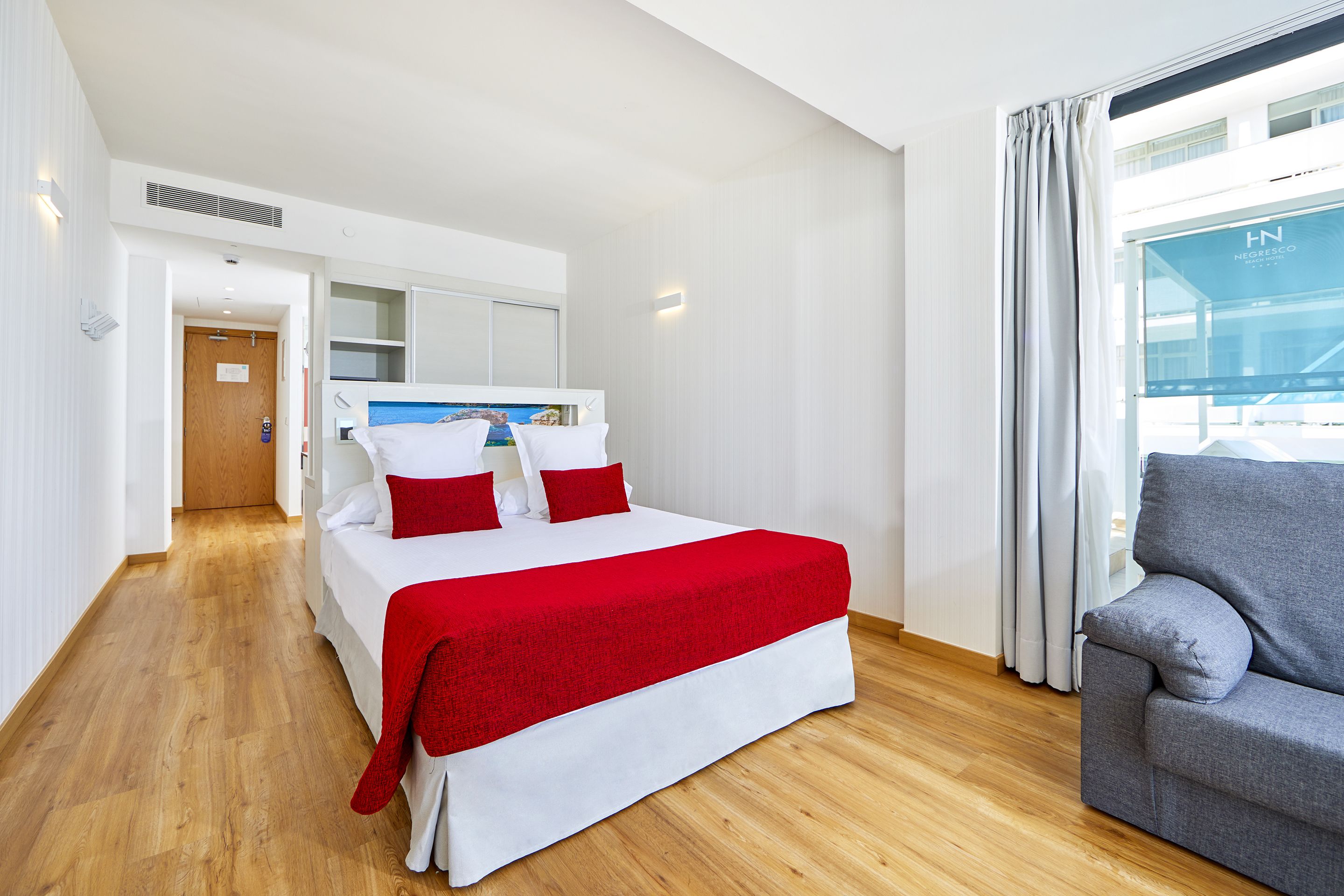 Ein Hotelzimmer mit einem Bett, einem grauen Sofa, Holzboden, einem Fenster mit Vorhängen und Blick auf ein Gebäude.