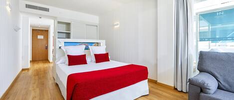 Camera d'albergo con un letto, un divano grigio, pavimento in legno, una finestra con tende e vista su un edificio.