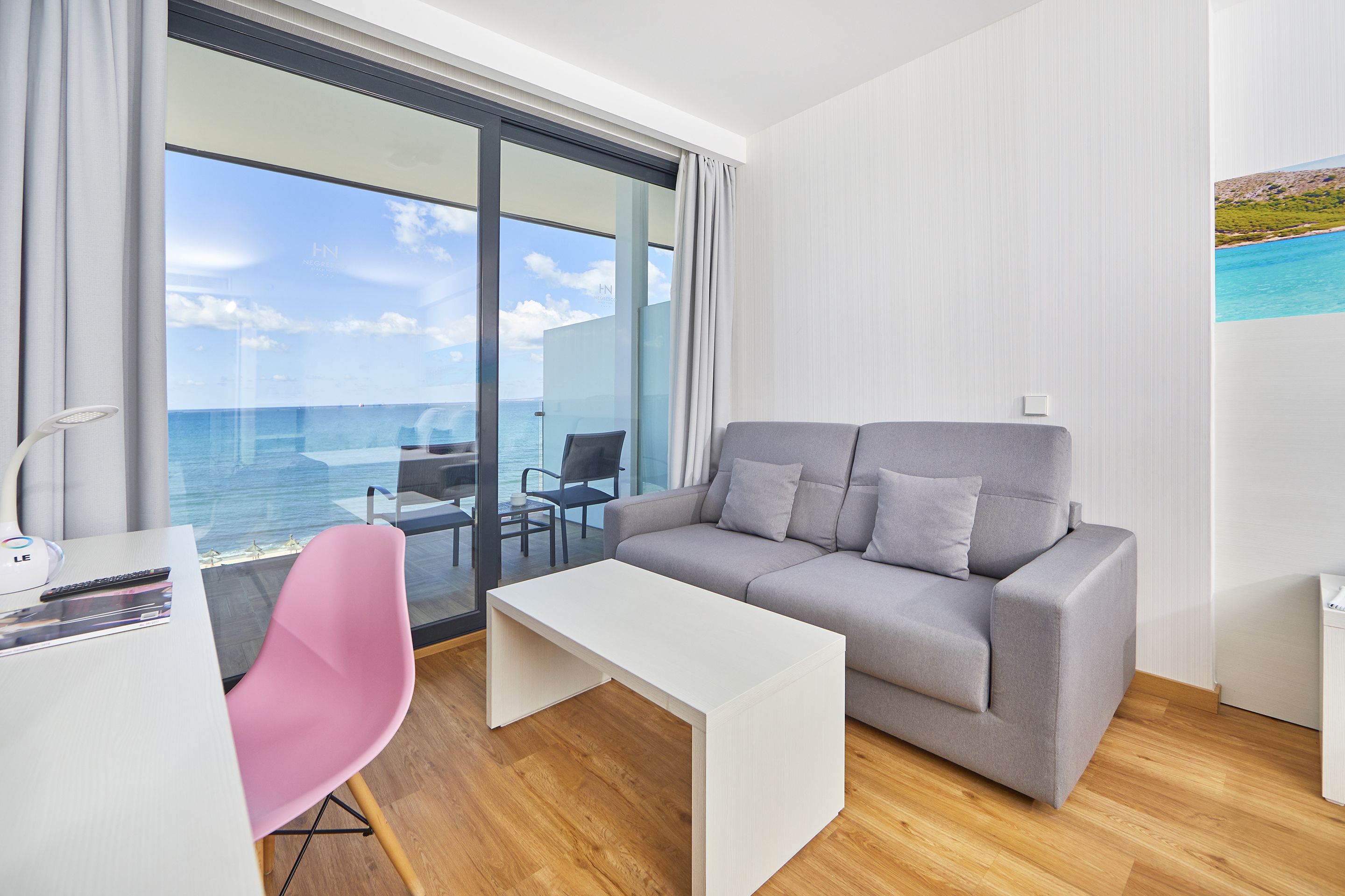 A modern hotel room with a grey sofa, white coffee table, and a pink chair. A large window offers a view of the sea.