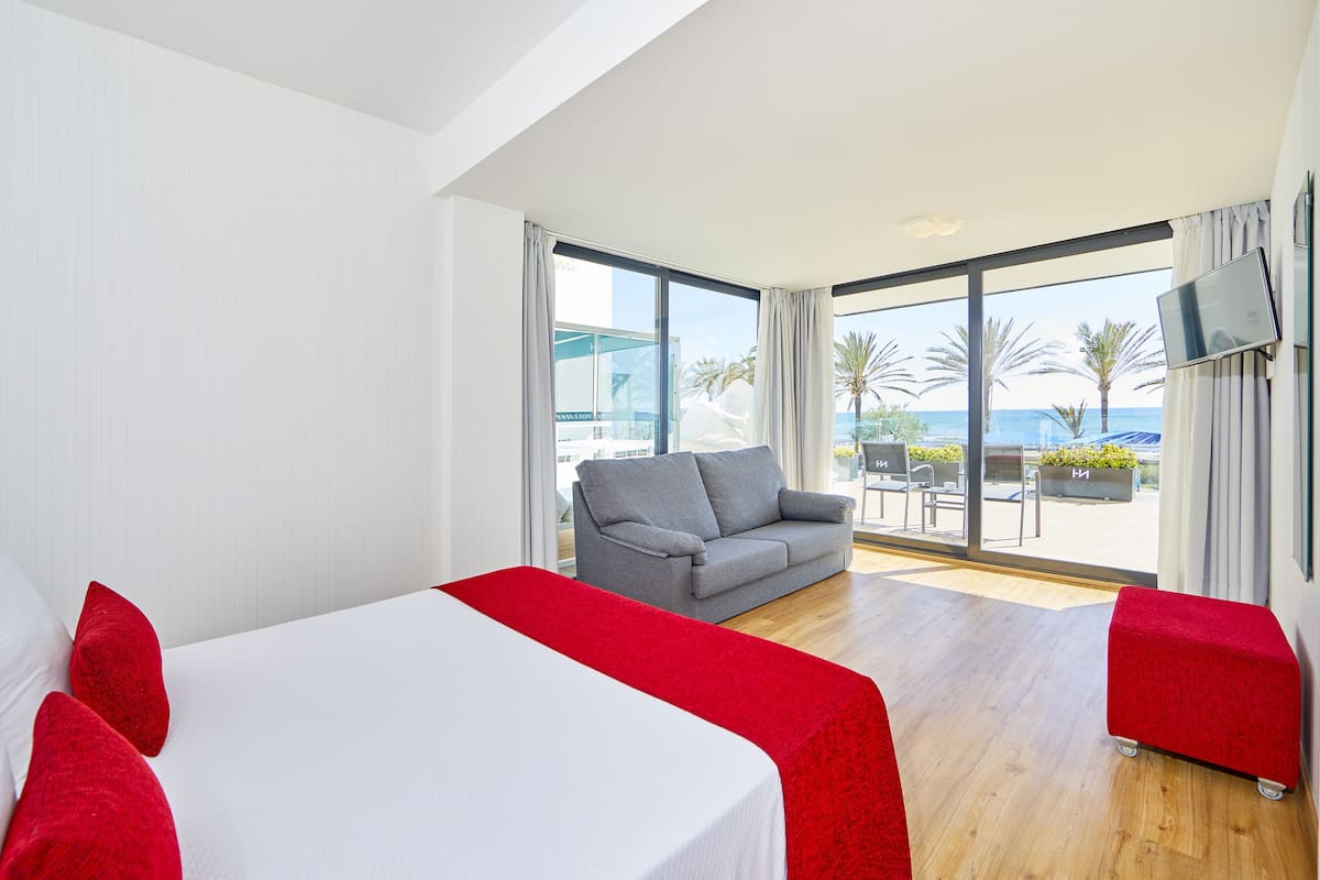 Habitación de hotel con una cama, un sofá gris y un reposapiés rojo. Se ve un ventanal con vistas a palmeras y al mar.