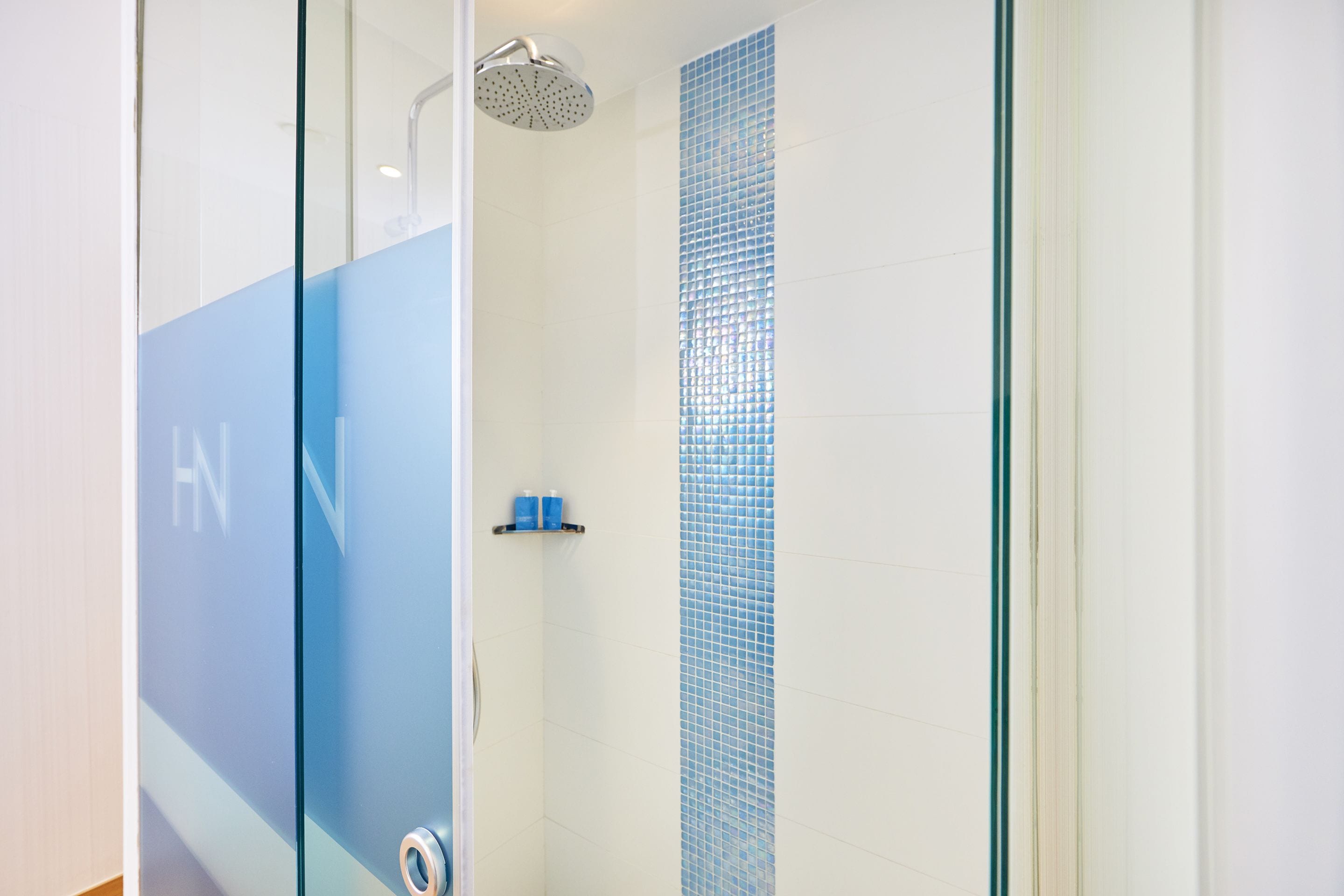 A modern shower with a glass door, a blue-tiled wall, and a shelf with toiletries.
