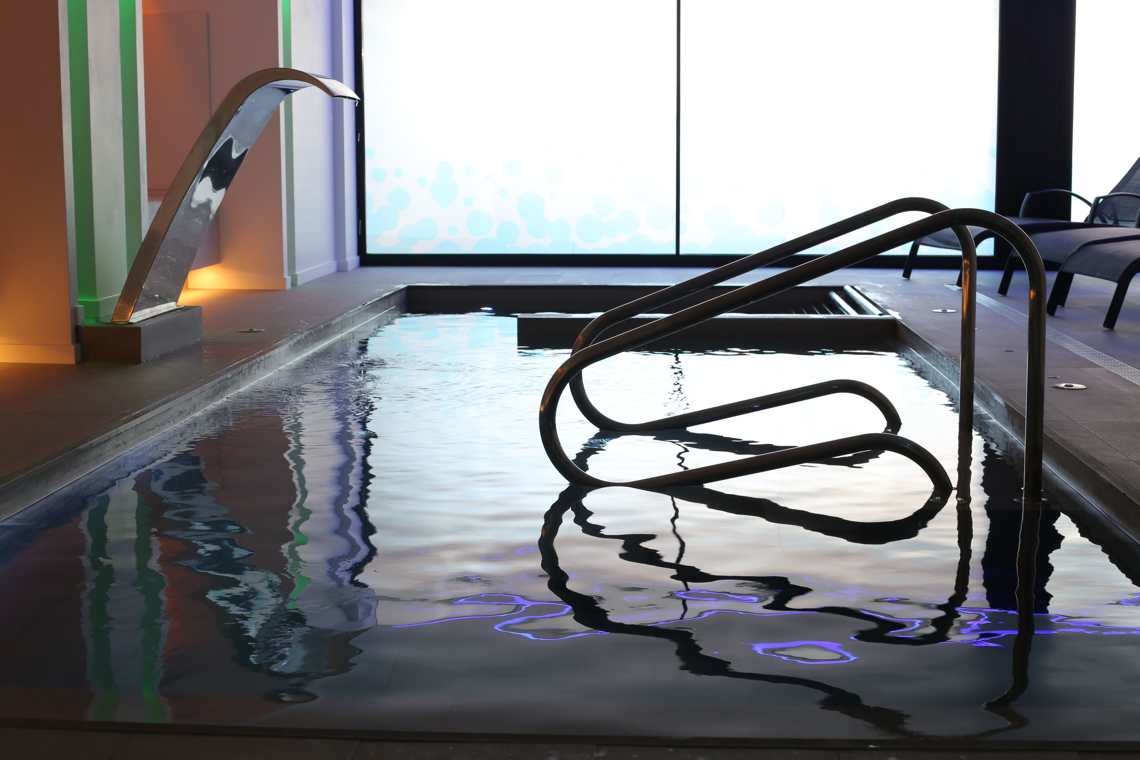 Una piscina con una escalera de metal y una pared de vidrio al fondo.