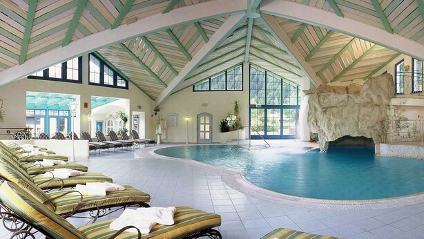 An indoor swimming pool with lounge chairs, a rock feature, and a high ceiling with green beams. - Alpenrose - Wellnessresort (Elbigenalp)