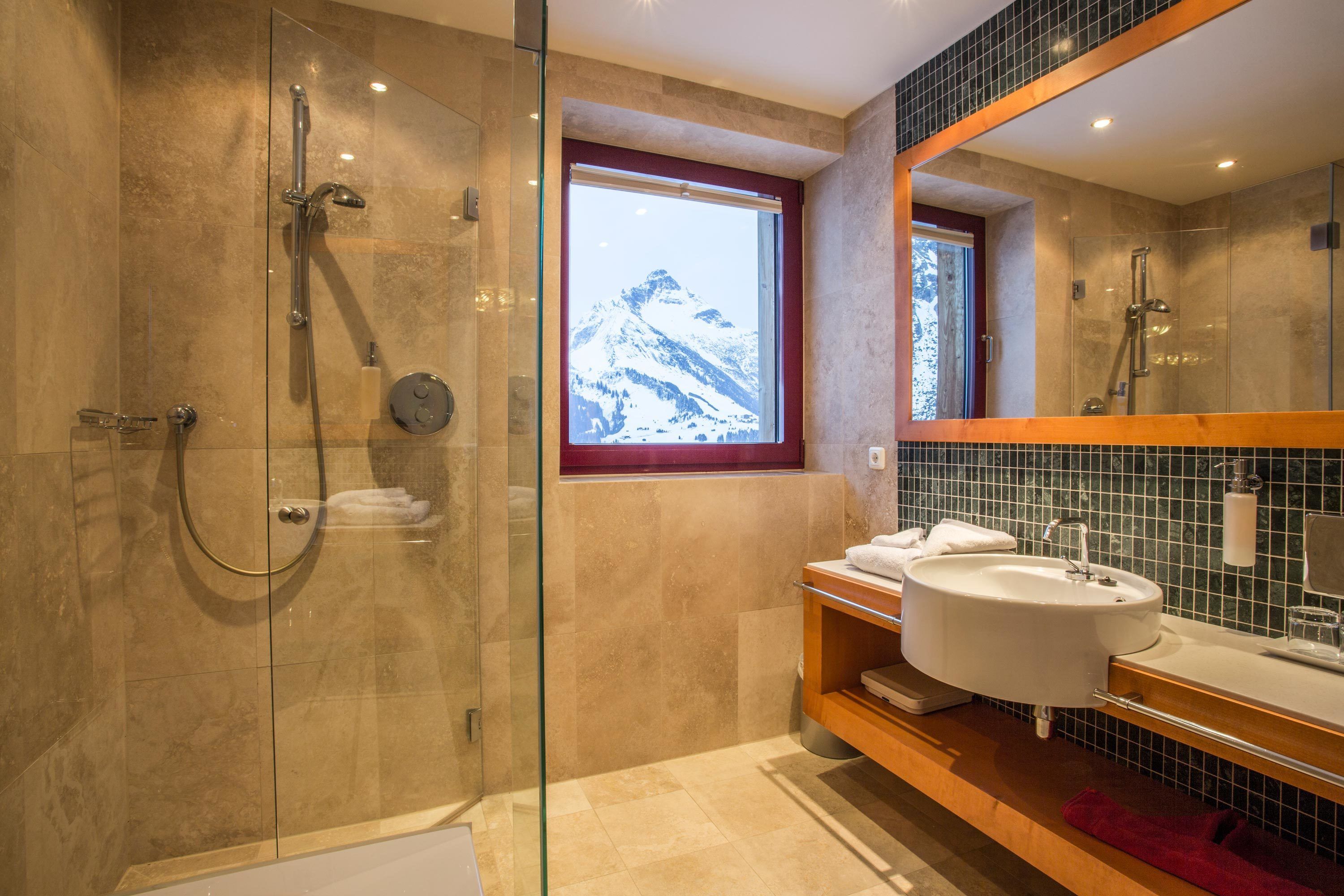 Un baño moderno con ducha de vidrio, un espejo grande y vista a una montaña nevada a través de una ventana.