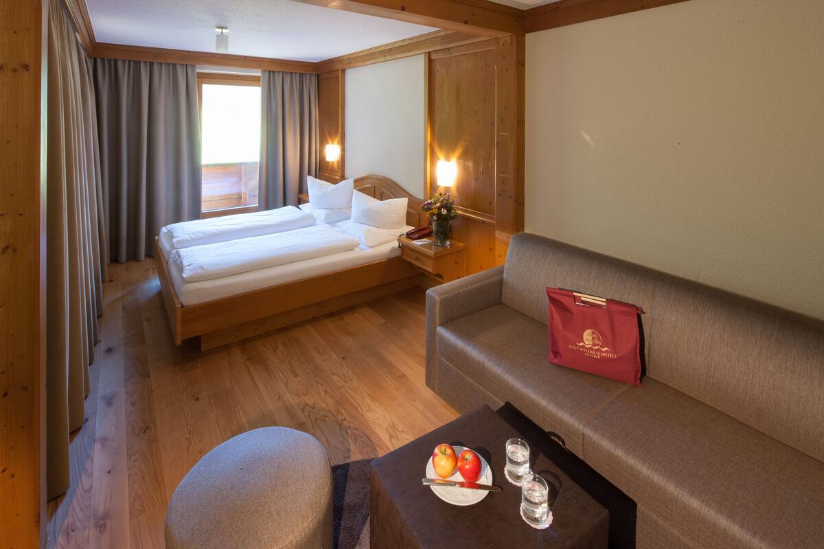 A hotel room with a bed, a sofa, a small table with apples and glasses, and a red bag on the sofa.