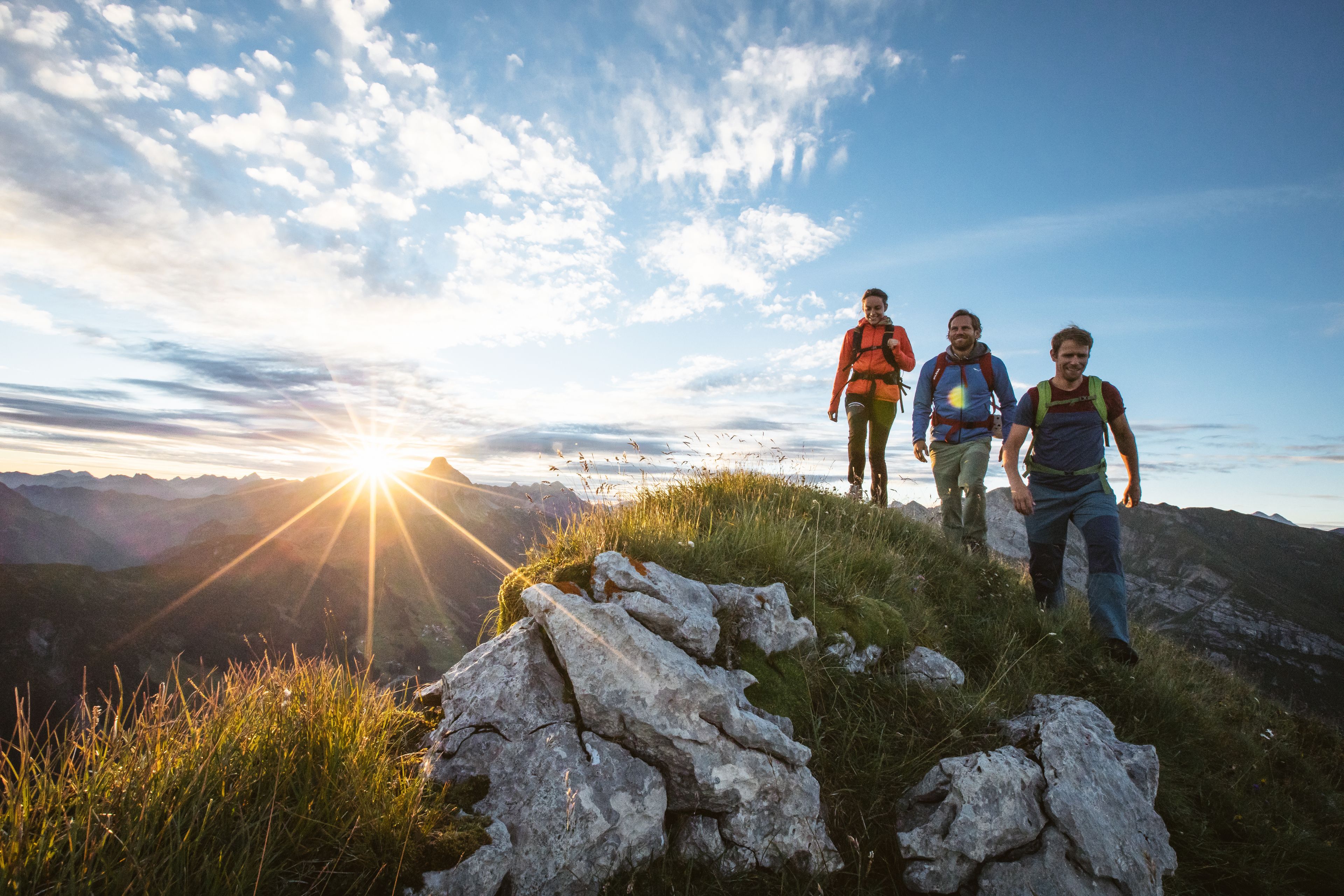 Drie wandelaars op een bergpad met een ondergaande zon op de achtergrond.