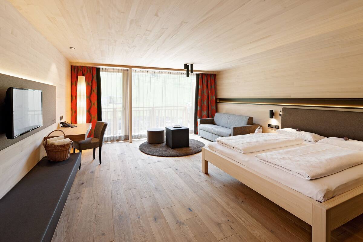 A modern hotel room with a wooden floor, a bed with white linens, a sofa, a desk with a chair, a television, and a large window with curtains.