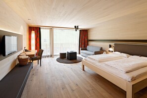 Una habitación de hotel moderna con suelo de madera, una cama con ropa blanca, un sofá, un escritorio con silla, un televisor y un gran ventanal con cortinas.