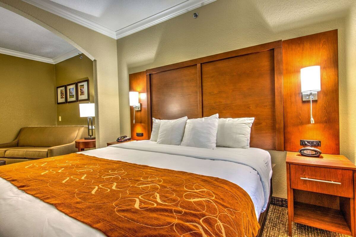 Une chambre d’hôtel avec un grand lit, une tête de lit en bois, deux tables de chevet, une petite table et une lampe fixée au mur.