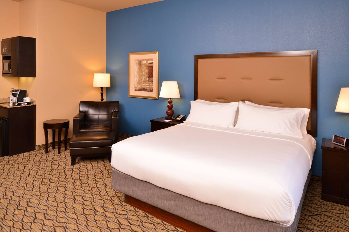A hotel room with a large bed, a chair, a desk with a lamp, and a painting on the wall.