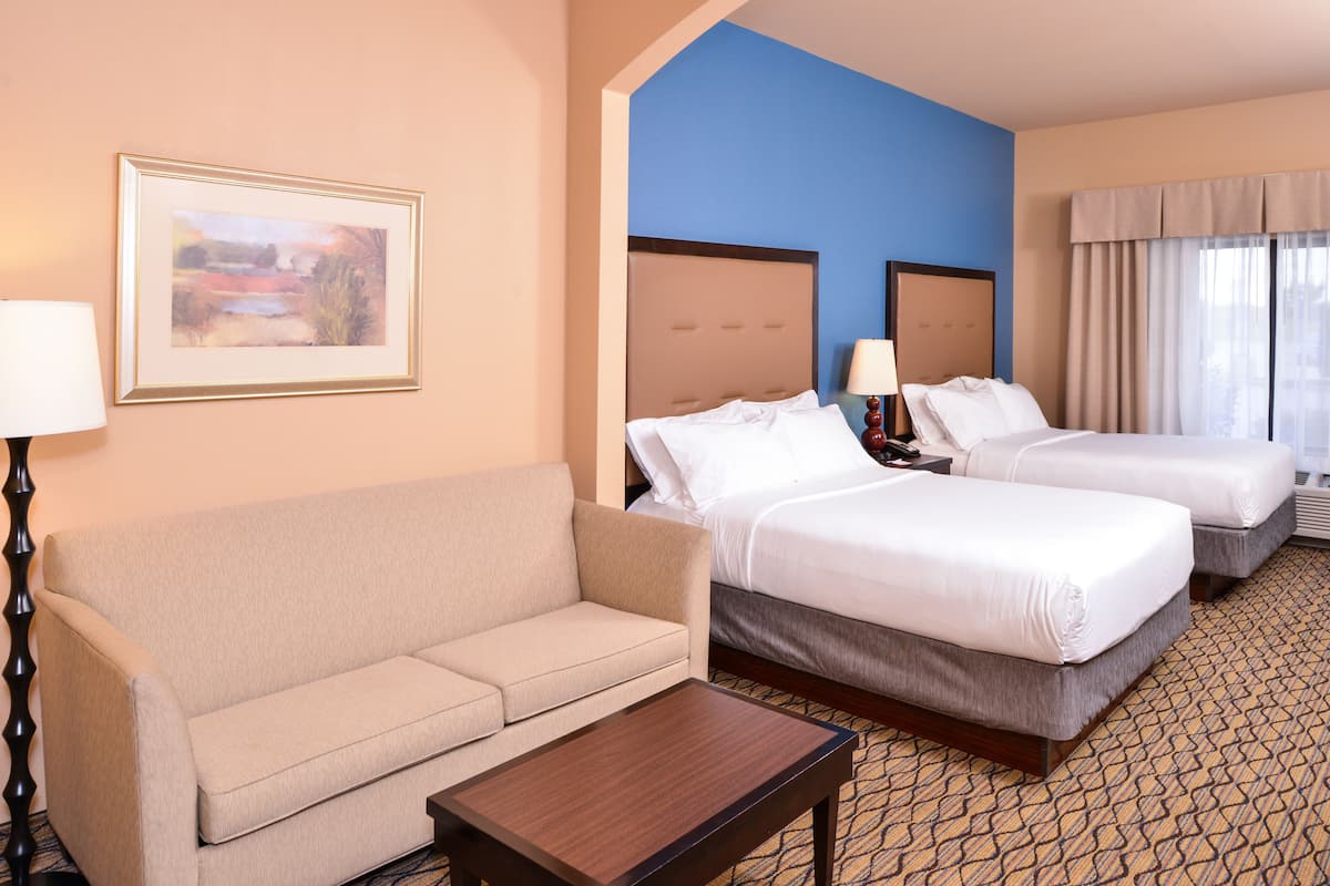 A hotel room with a sofa, two beds, a framed picture on the wall, and a lamp.