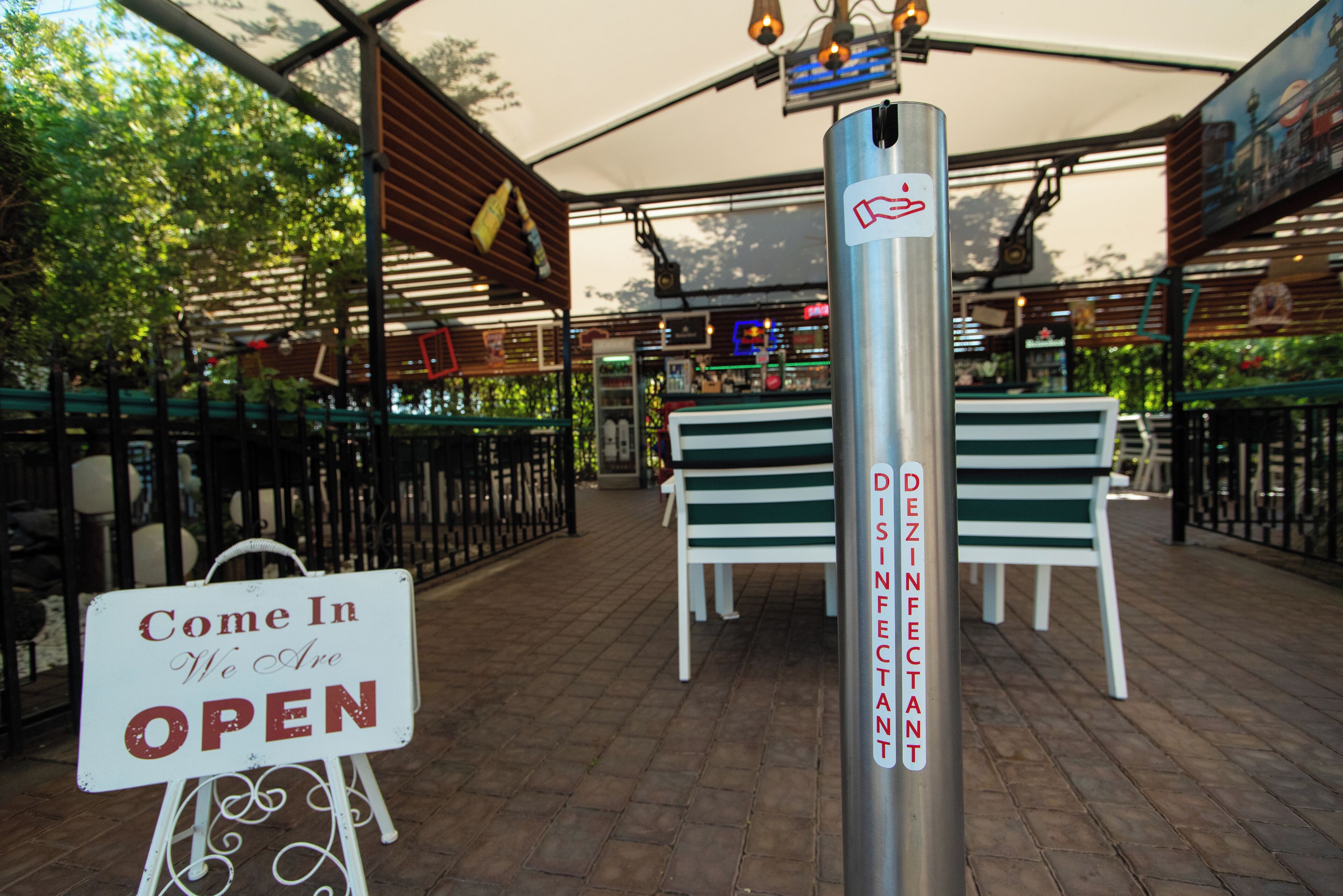 Un espacio al aire libre con asientos, un letrero que dice 'Come In We Are OPEN', bancos verdes y una estación dispensadora de desinfectante.
