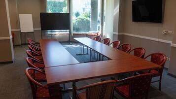 Ein Konferenzraum mit langem Tisch, roten Stühlen, einer Whiteboard-Tafel, einem Bildschirm und einem Fernseher an der Wand.