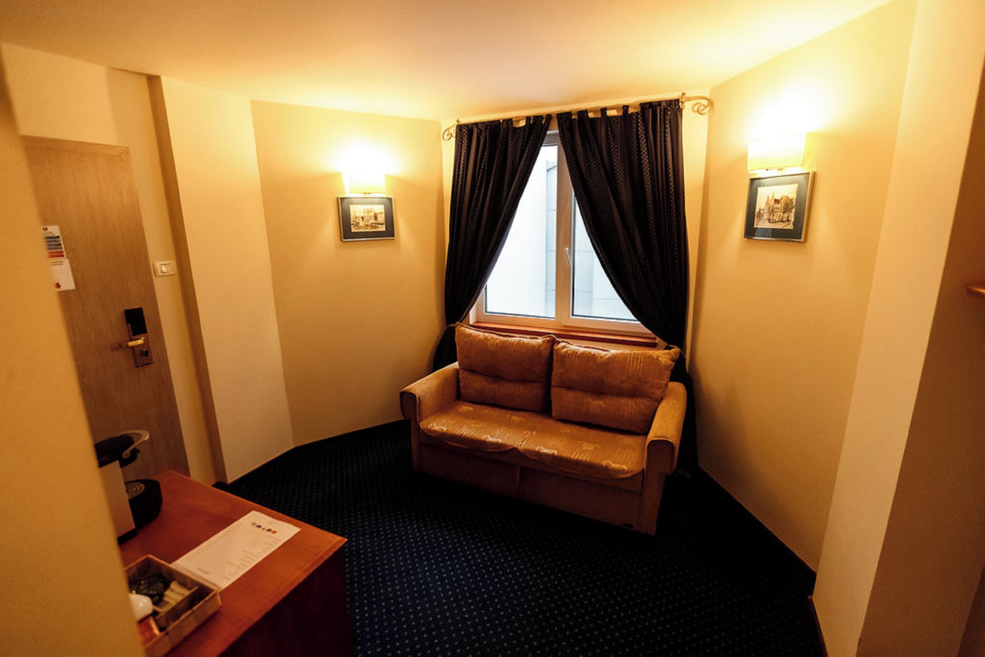 A hotel room with a brown leather sofa, a desk with a menu, two wall-mounted lamps, and a window with curtains.