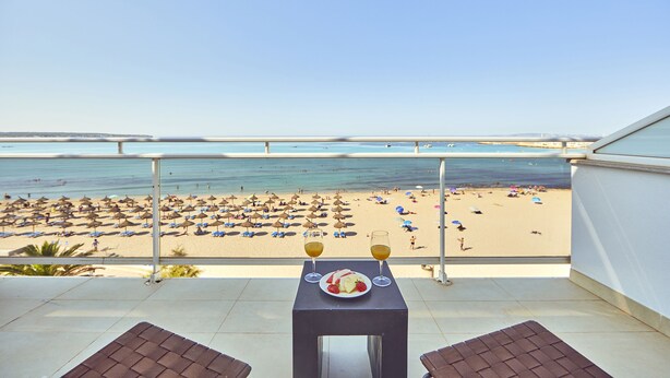 Un balcon avec une table dressée pour deux personnes, donnant sur une plage parsemée de parasols et animée par la présence de vacanciers.