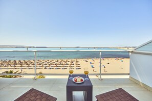 Ein Balkon mit Tisch für zwei Personen, mit Blick auf einen Strand mit Sonnenschirmen und Menschen.
