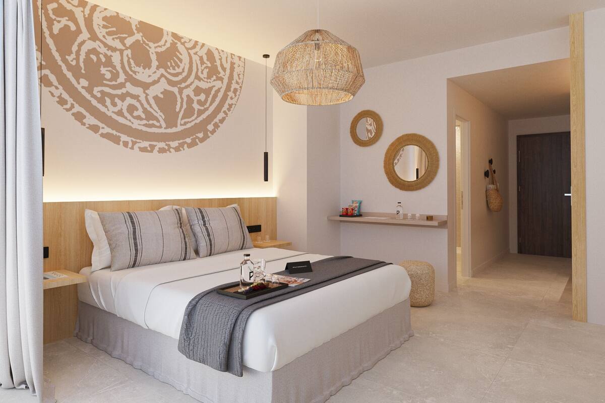 A modern bedroom with a large bed, bedside tables, a hanging light fixture, a wall-mounted mirror, and a door leading to another room.