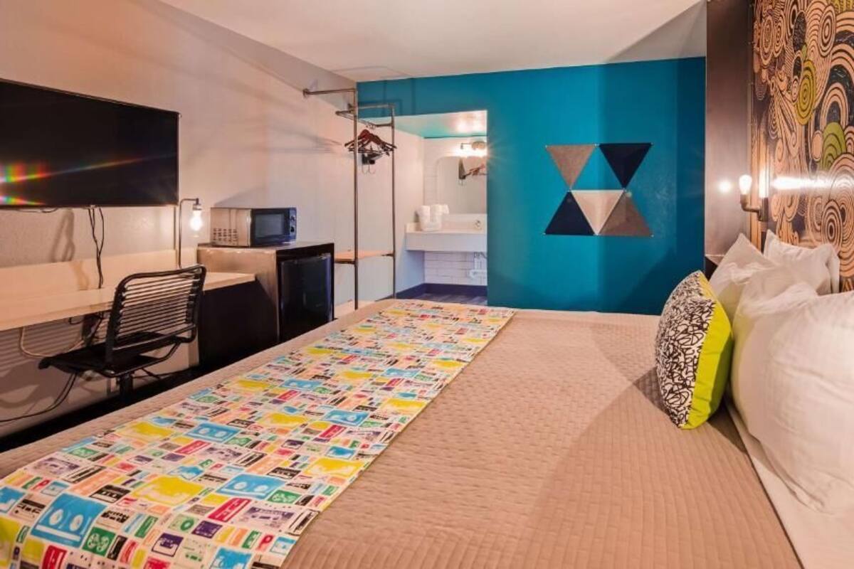 A hotel room with a bed, a desk, a chair, a TV, and a bathroom visible through an open door.