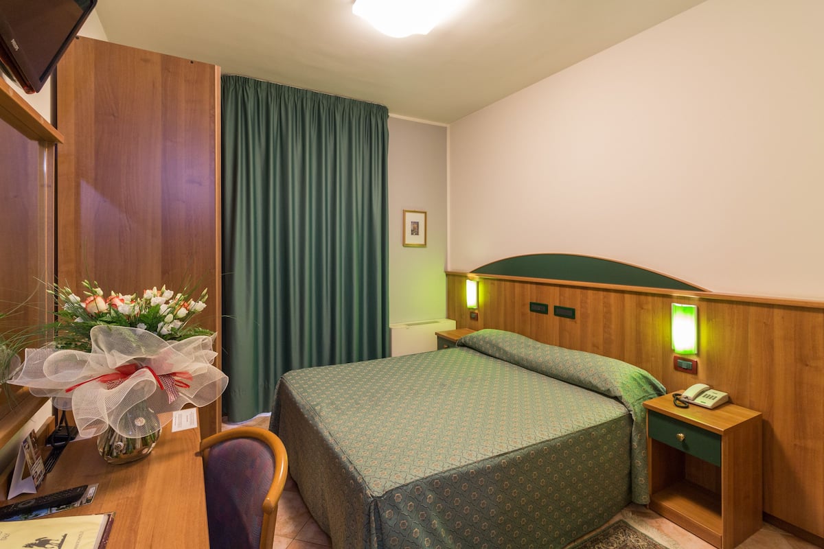 Una habitación de hotel con cama, escritorio con teléfono, silla, lámpara de escritorio, mesita de noche con lámpara y cortina.