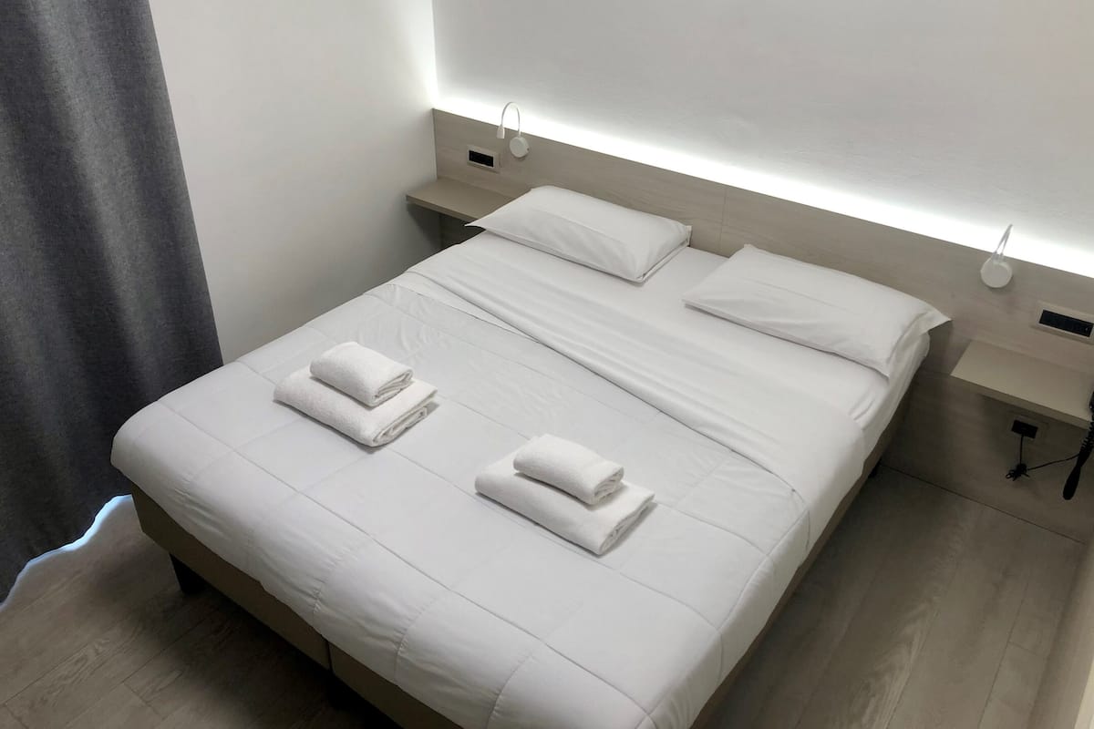 Una cama bien hecha con sábanas blancas y dos toallas dobladas, una mesita de noche con lámpara, y un aplique de pared.