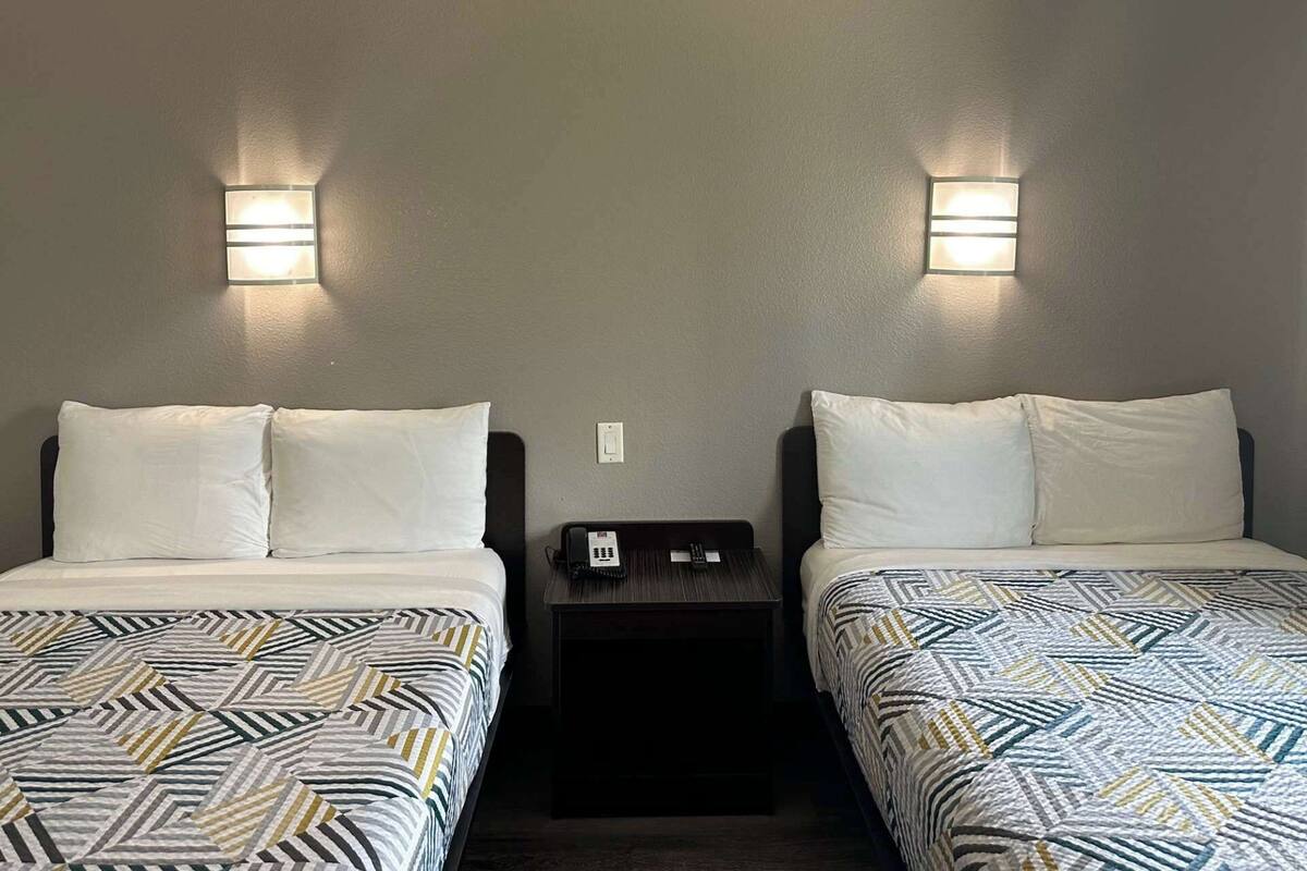 A hotel room with two beds, each with a patterned bedspread, a nightstand with a phone, and wall-mounted lights.