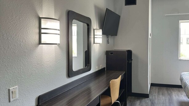 A compact hotel room with a dark wood desk, a chair, a large mirror, a flat-screen TV mounted on the wall, and a door leading to another room. - Motel 6 Mission, TX (Mission)