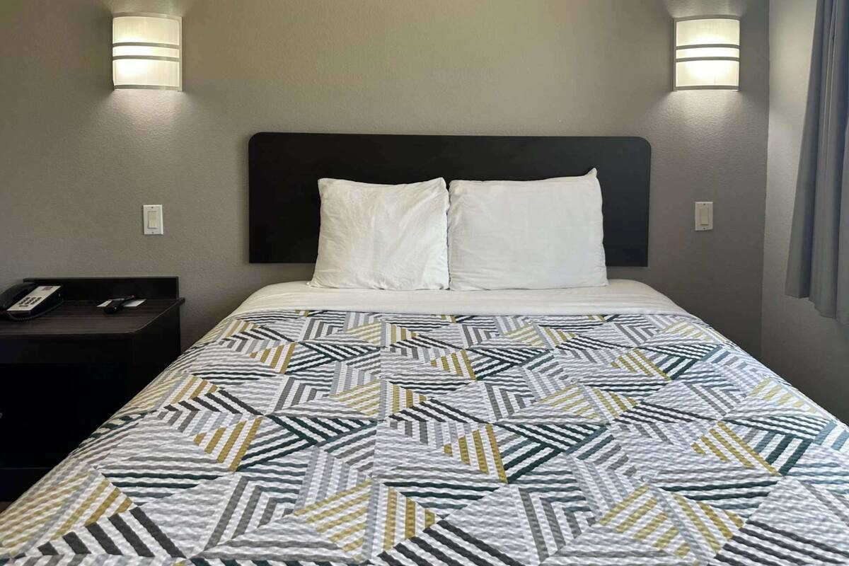 A hotel room with a bed, two bedside lamps, a nightstand with a phone, and a patterned bedspread.