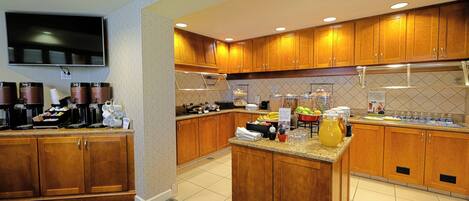 A hotel kitchen with wooden cabinets, a flat-screen TV, and various breakfast items on the counter.