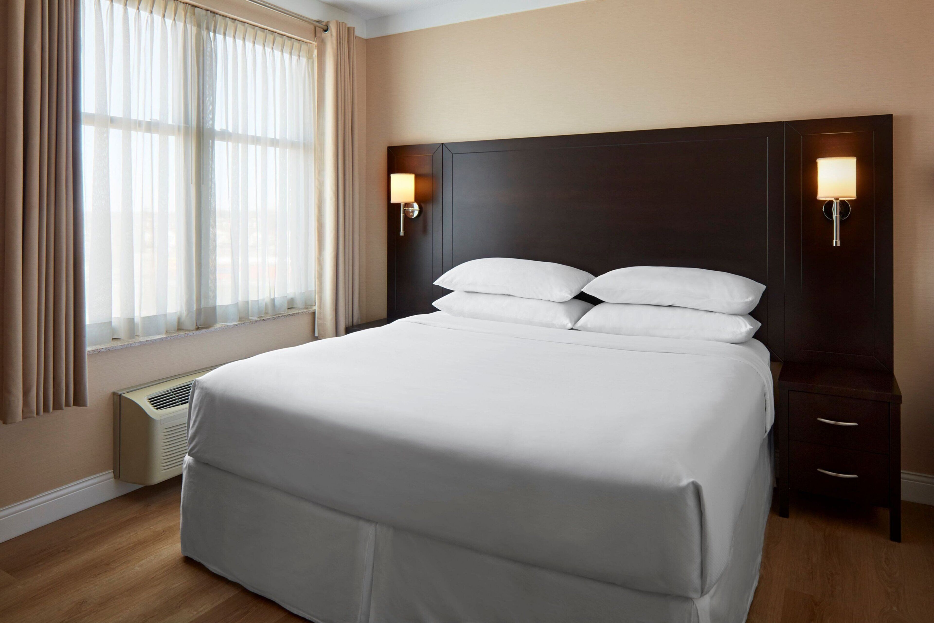 Una habitación de hotel con una cama grande, dos mesitas de noche con lámparas, una ventana con cortinas y un calentador fijado a la pared.