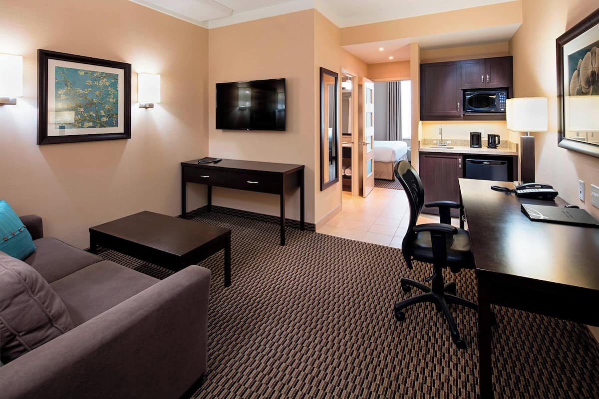 A hotel room with a sofa, a desk, a TV, a kitchen area with a microwave, and a dining table.