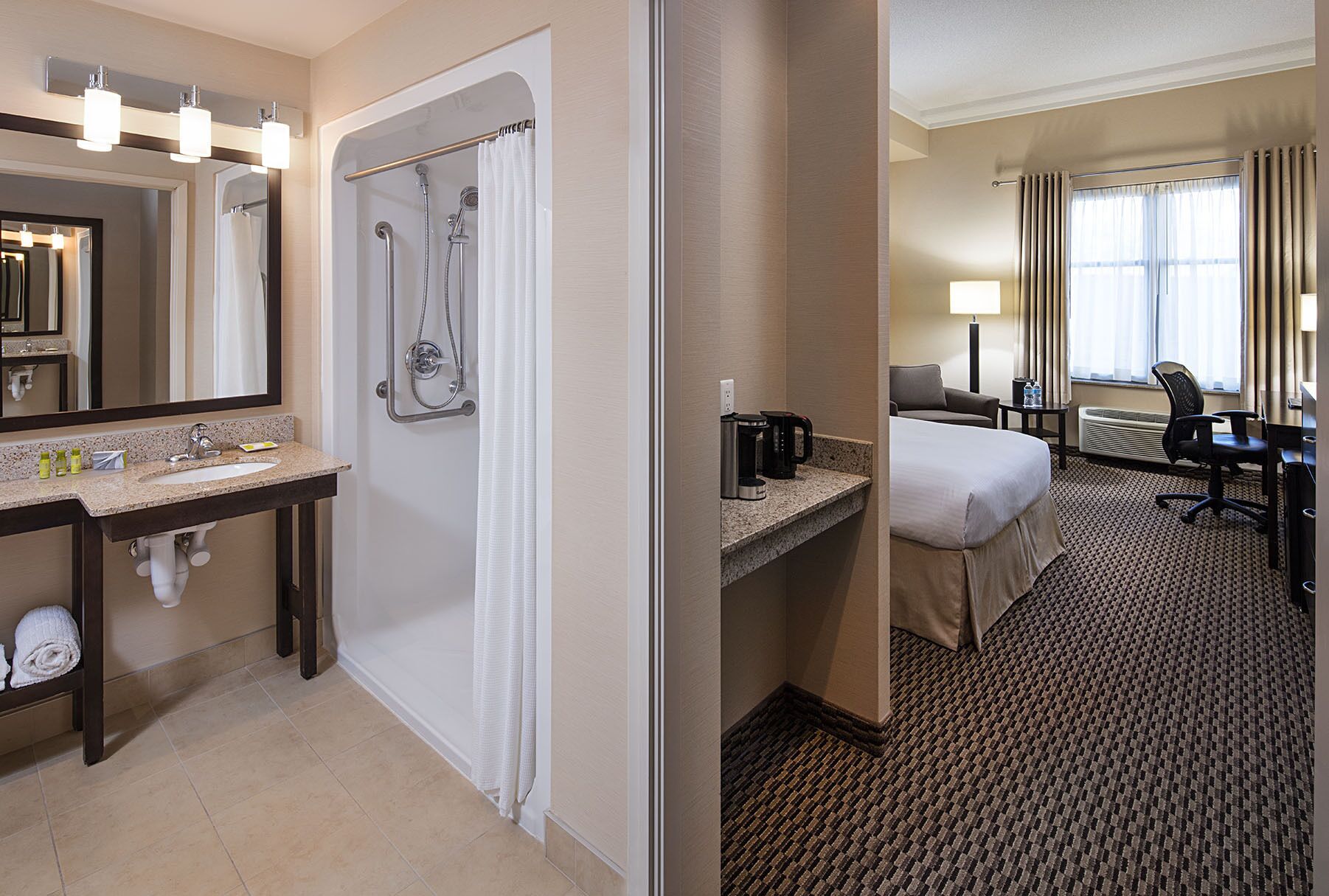 Un baño de hotel con lavamanos, espejo, ducha y una cama en la habitación contigua.