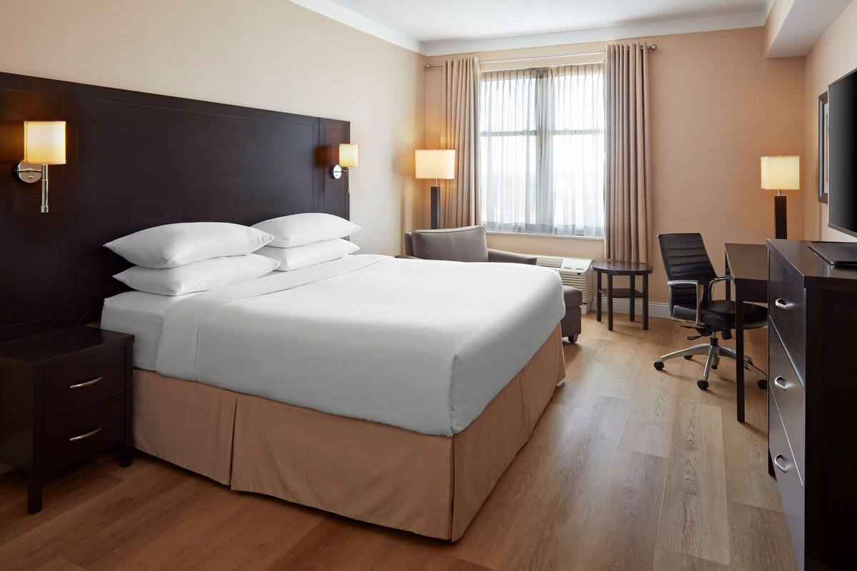A hotel room with a large bed, bedside tables, a desk with a chair, a TV, and a window with curtains.