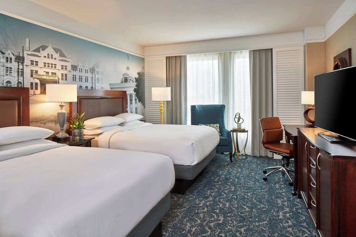 A hotel room with two beds, a desk, a TV, and a mural of a building.