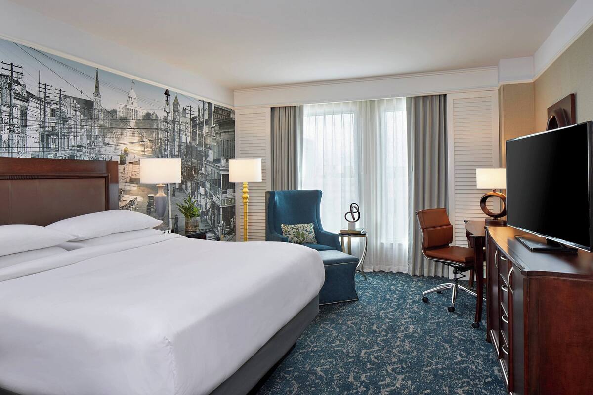 A hotel room with a large bed, a desk with a TV, a chair, and a desk lamp.